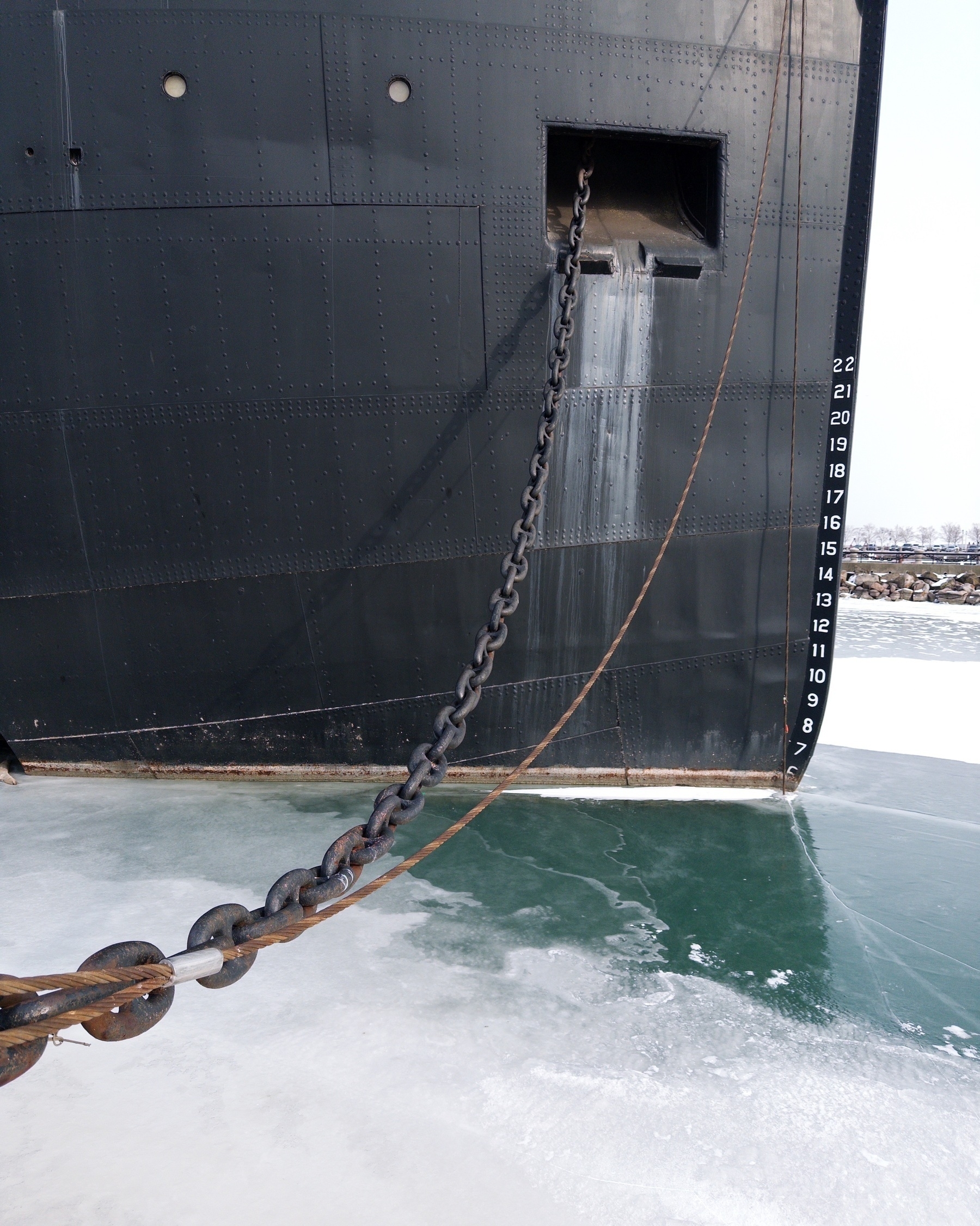 A large ship's black hull is anchored with a heavy chain into a frozen body of water.