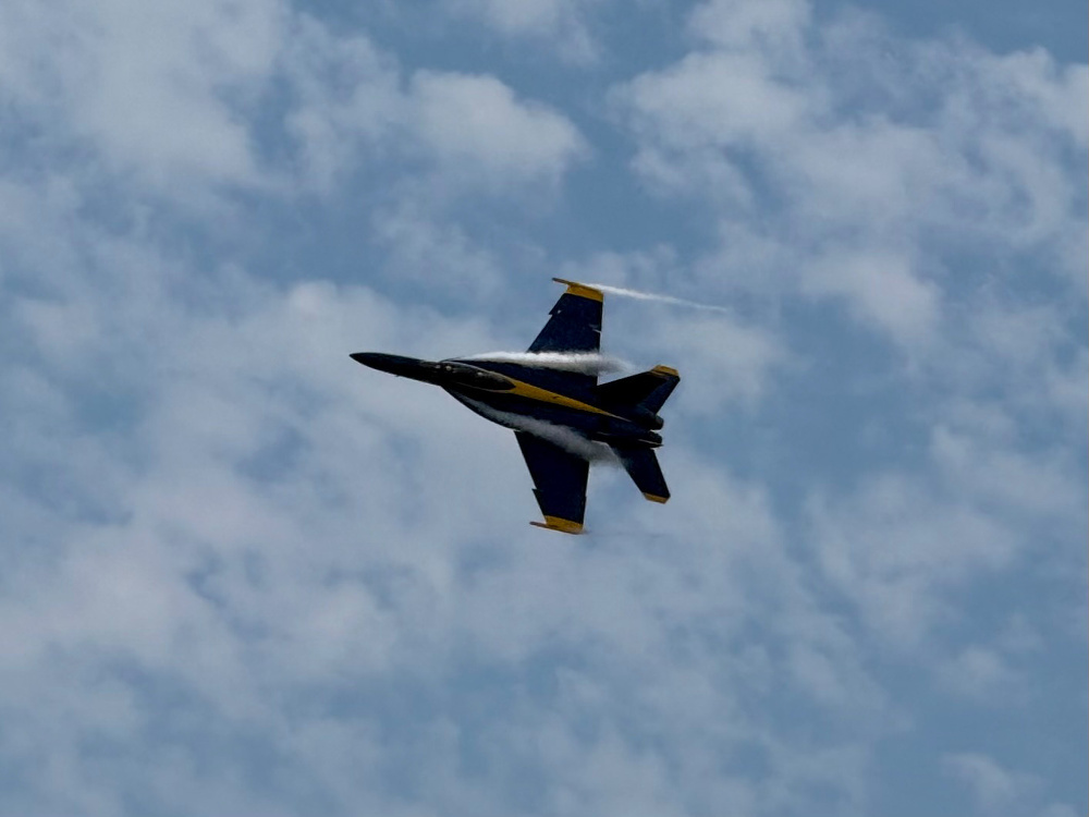 A jet fighter with blue and yellow markings is flying through a partly cloudy sky.