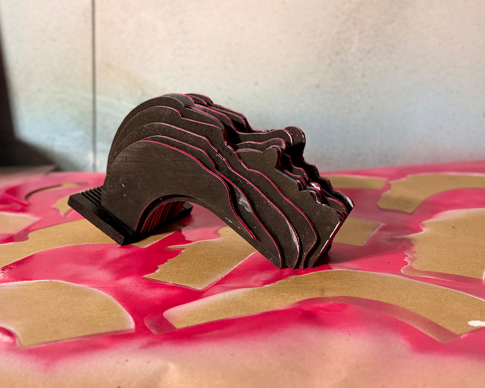 A multi-layered sculptural head profile made of black material with pink edges is placed on a surface covered in brown paper and pink paint or spray.