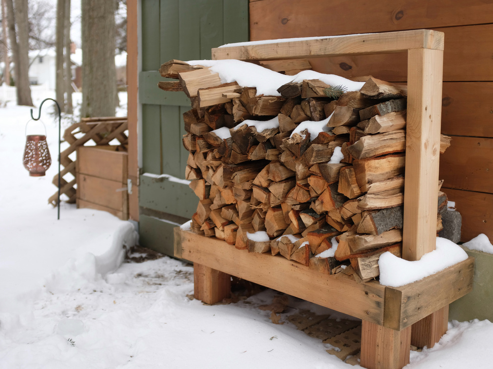 A stack of firewood covered with snow is neatly arranged on a wooden rack beside a building in a snowy outdoor setting.