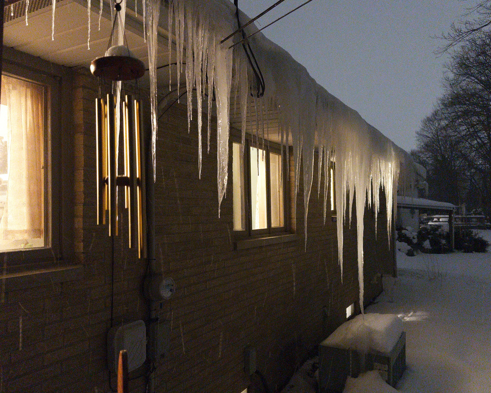 Large icicles hang from the edge of a roof on a snowy winter evening.
