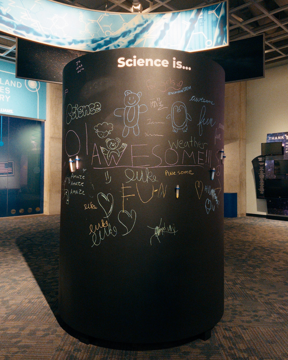 A cylindrical blackboard with the heading Science is... is covered in colorful chalk drawings and positive words like awesome, fun, and weather inside a museum or science center.