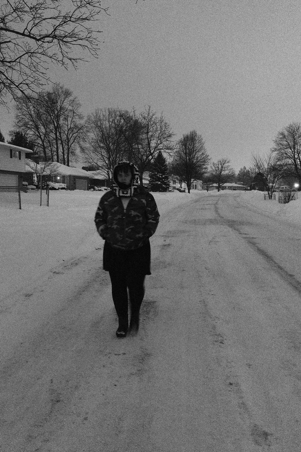 A person wearing a camouflage jacket walks down a snowy street lined with trees and houses in a black and white scene.