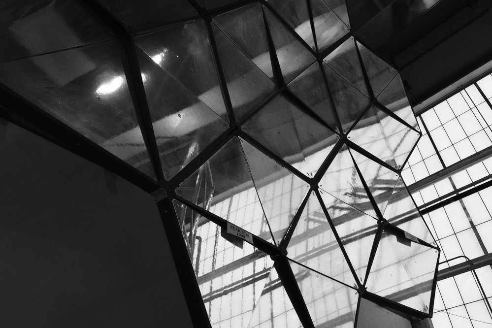 A geometric sculpture composed of transparent triangular panels stands in front of large industrial windows.