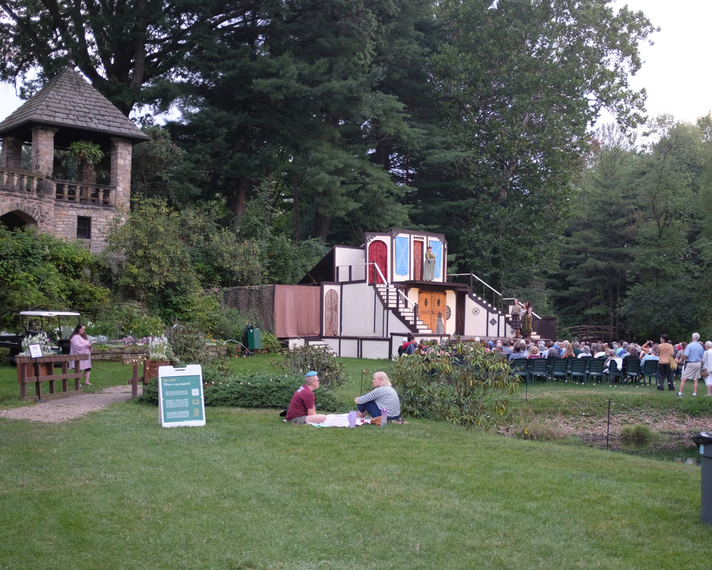 A small outdoor theater set is surrounded by people, some seated in chairs and others on the grass, with a brick gazebo and lush trees in the background.