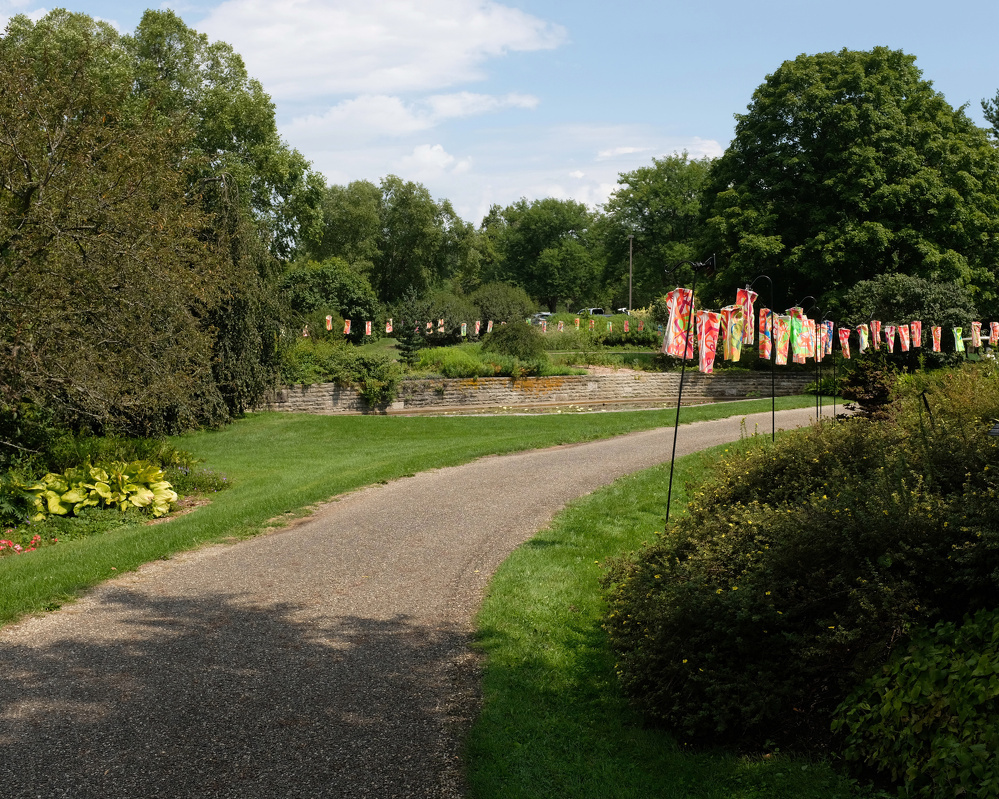 A winding pathway through a green park is lined with colorful hanging decorations on poles.