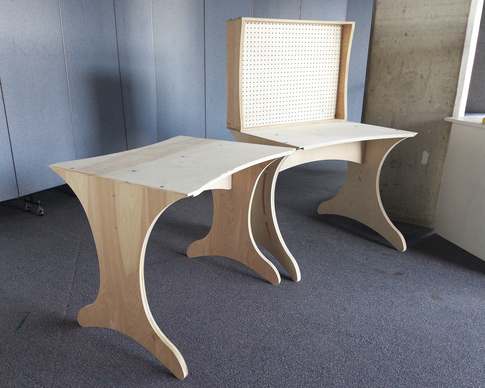Two wooden tables with curved legs are positioned in a room with blue walls and a carpeted floor, one of which has a pegboard attached to its back.