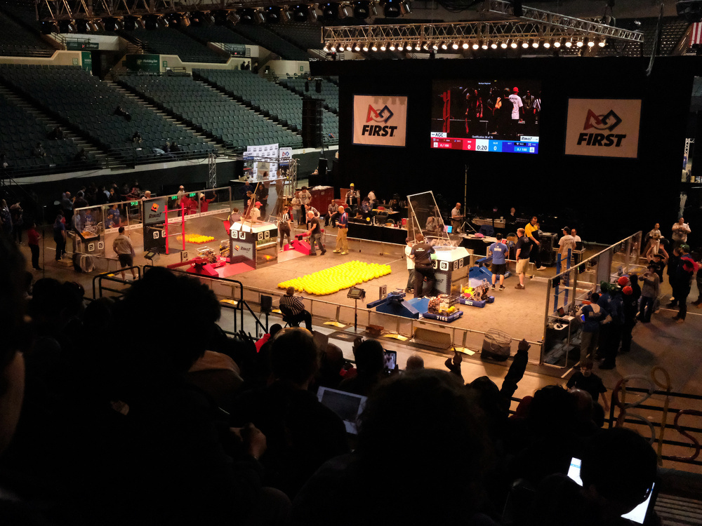 A robotics competition is taking place in an arena, with teams working on their robots and spectators watching from the stands.