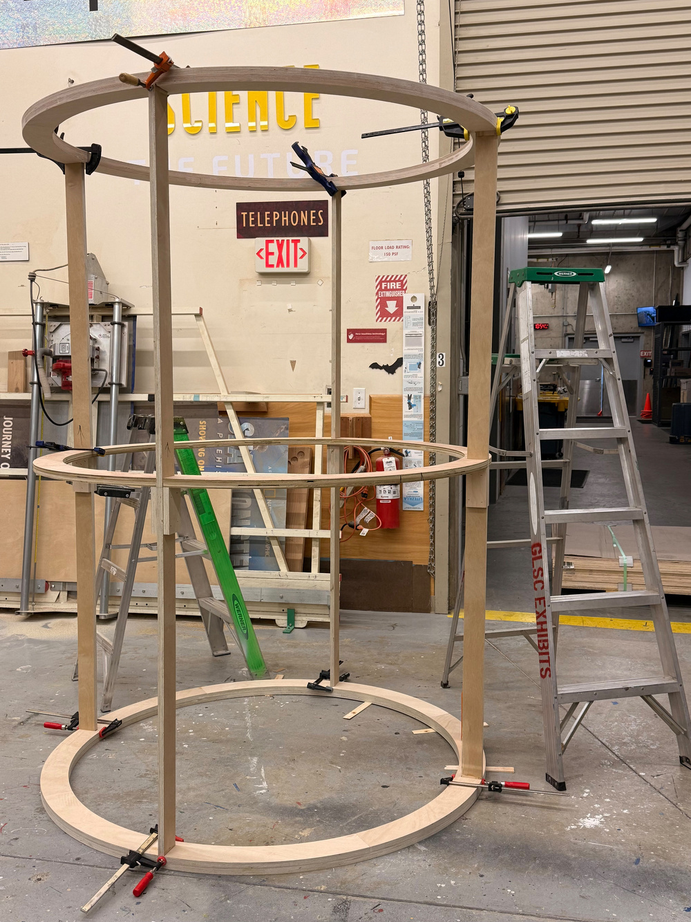 A partially constructed circular wooden frame is held together with clamps inside a workshop filled with ladders, tools, and various materials.
