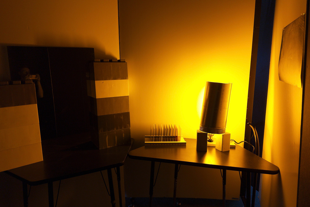 A warm yellow light illuminates a corner with tables holding several objects, including a black cylindrical item, a small white rack, and some box-like structures.