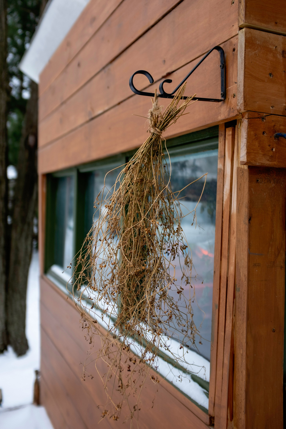 A bundle of dried plants hangs from a decorative hook on a wooden building exterior, with snow visible on the ground.