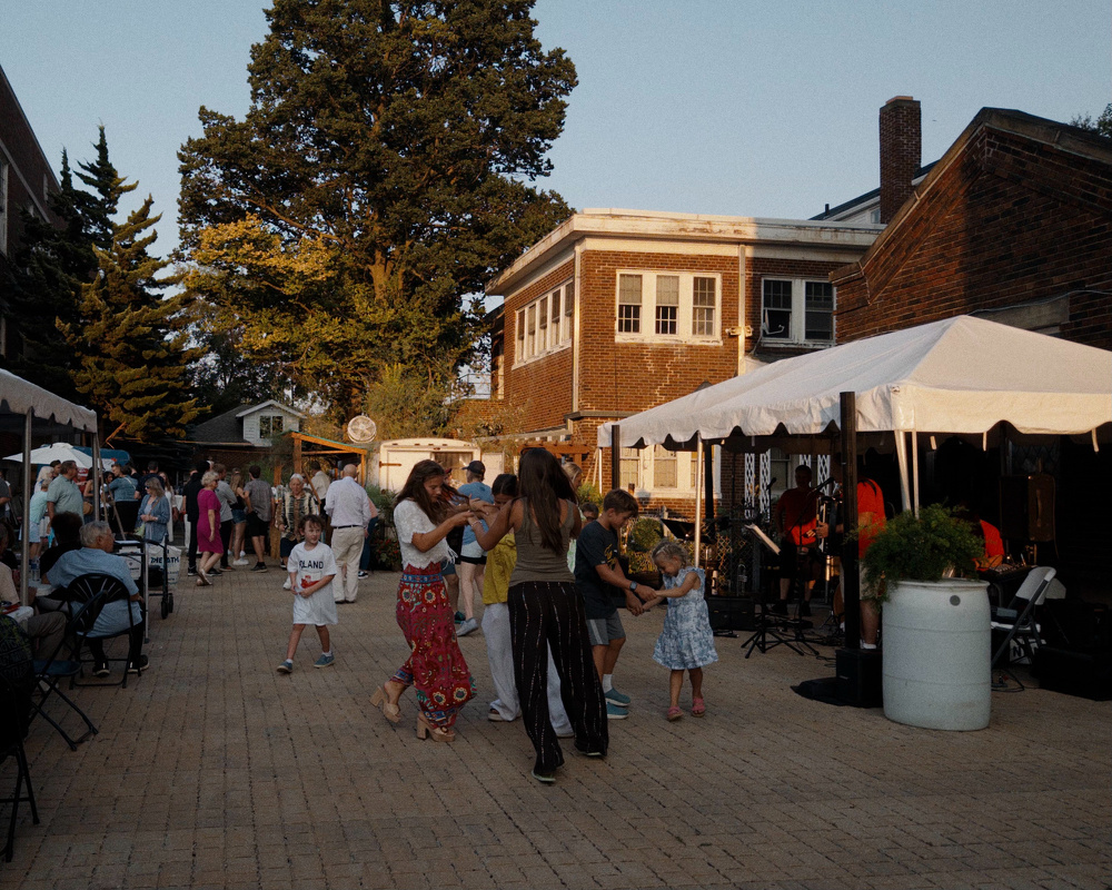 People are gathered in an outdoor setting with some dancing and others walking or sitting near tents and a brick building.