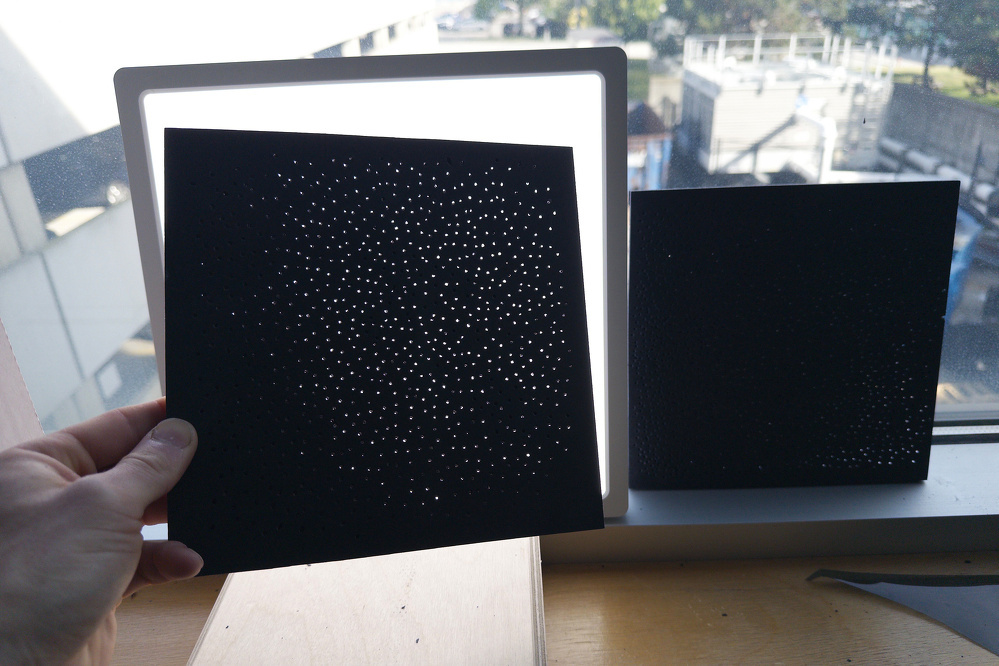 A person holds a perforated black sheet in front of a bright window, with another similar sheet leaning against the window ledge.