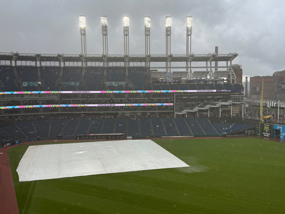 A baseball field is covered with a tarp while rain falls under overcast skies and stadium lights illuminate the scene.