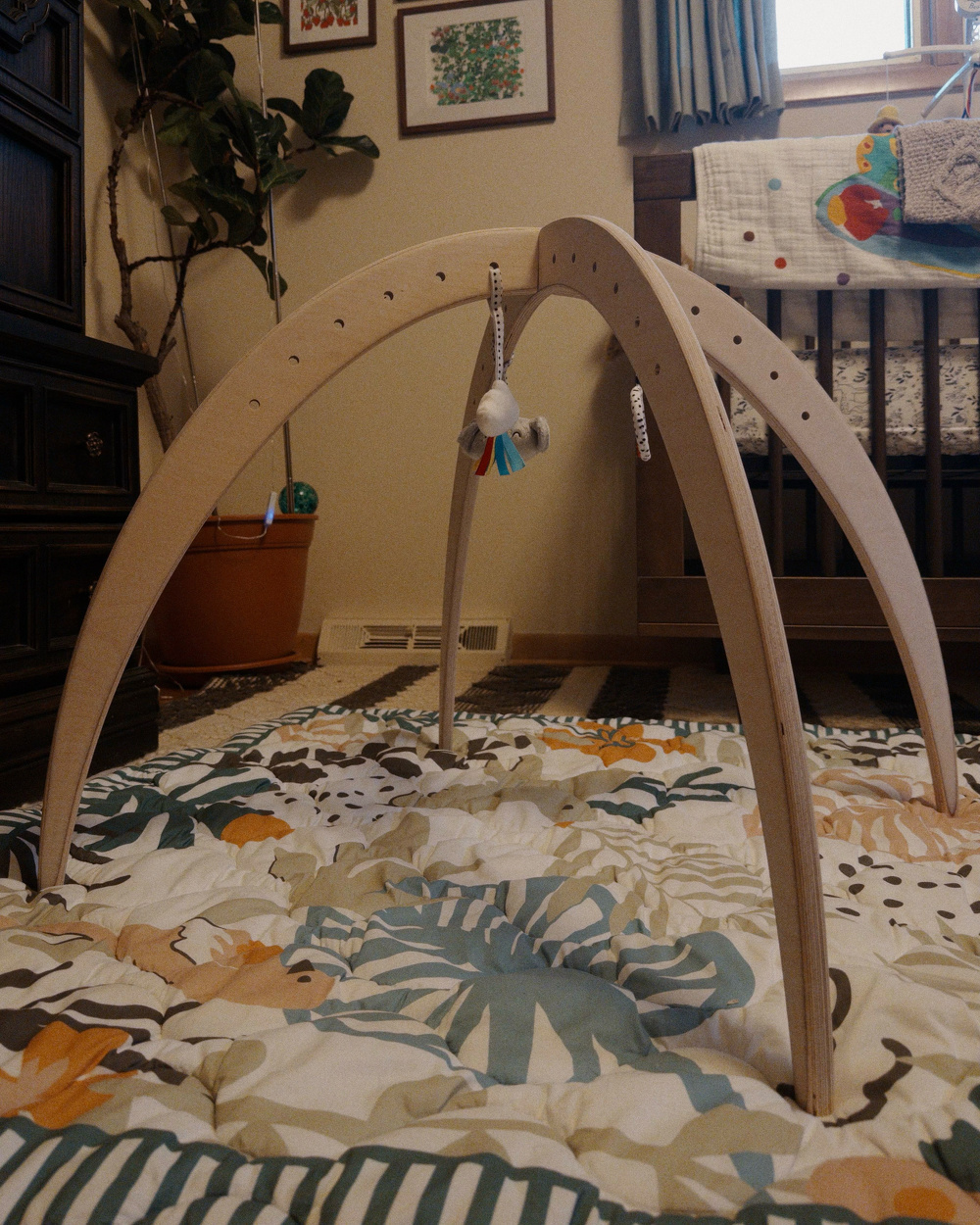 A wooden baby gym with hanging toys is set up on a colorful leafy-patterned quilt in a nursery room.