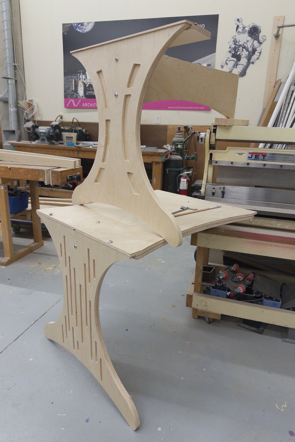 A uniquely designed wooden chair featuring curved and slotted structural elements is shown in a workshop environment.