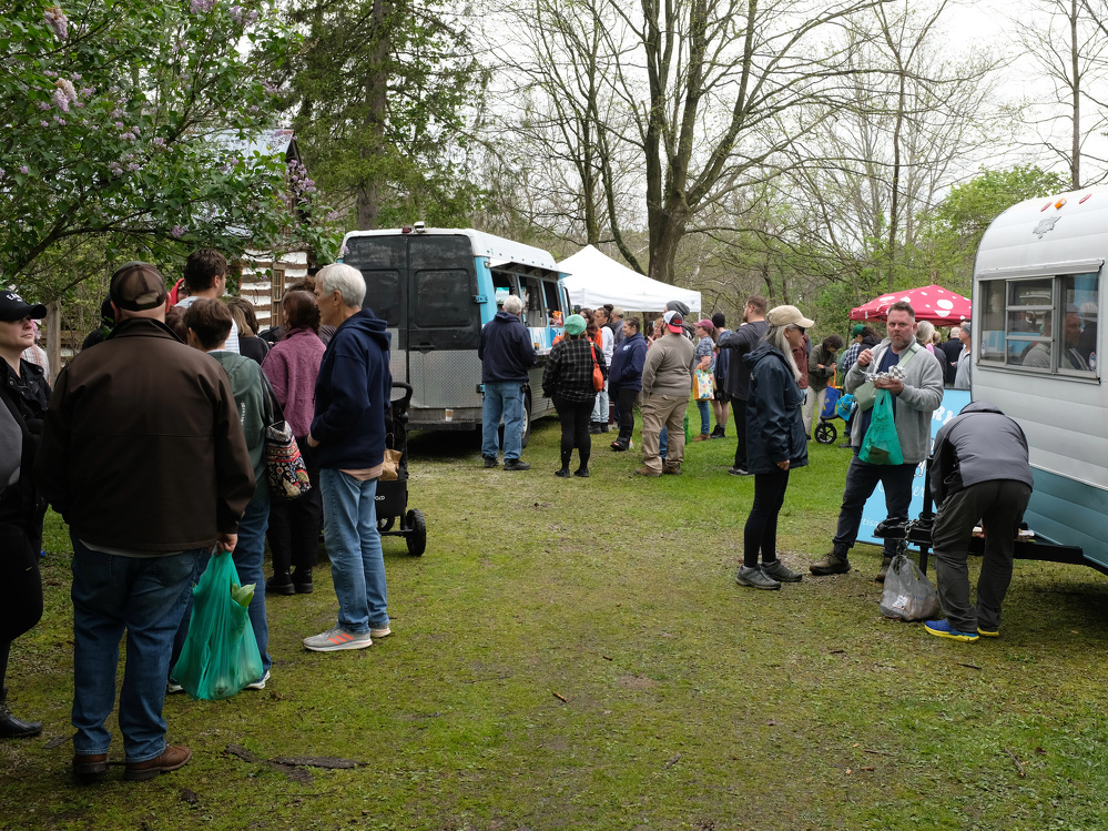 People are gathered around food trucks in a park setting with trees and tents nearby.