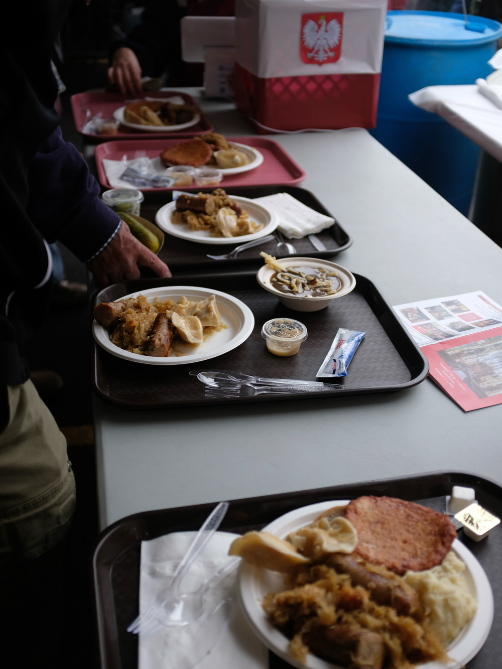 Several trays of food with Polish dishes and plastic utensils are lined up on a table, with a box featuring the Polish national emblem visible in the background.