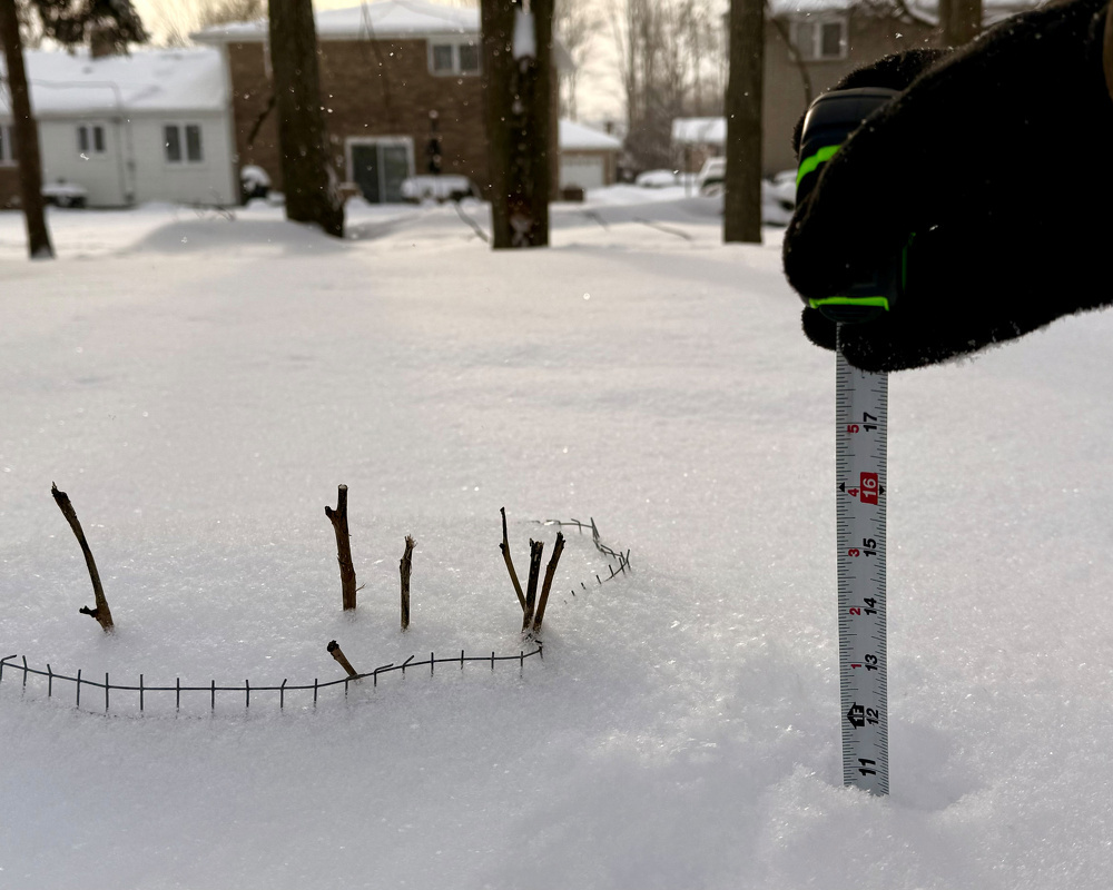 A hand in a glove is holding a measuring tape in the snow near some small twigs and a wire fence, with houses and trees in the snowy background.