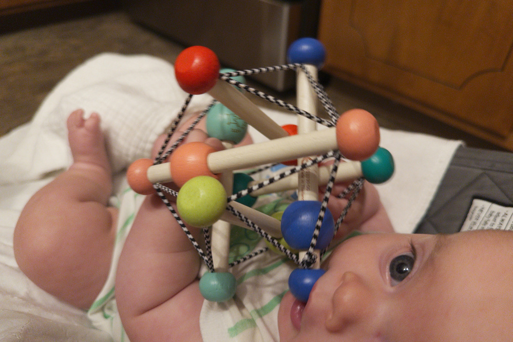 A baby is lying down and holding a colorful wooden toy with interconnected rods and balls.