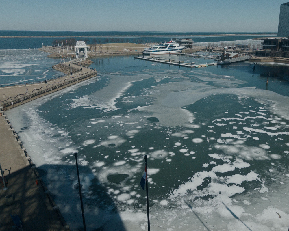 A partially frozen harbor with a docked boat and surrounding buildings is shown on a clear day.
