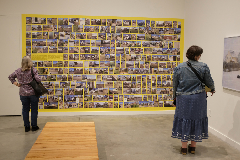 Two people observe a large collage of photographs displayed on a gallery wall.