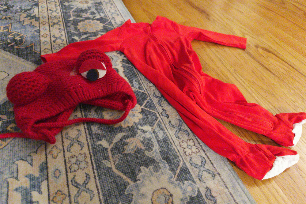 A red knitted hat with large eyes and a matching red onesie are laid out on a patterned rug and wooden floor.