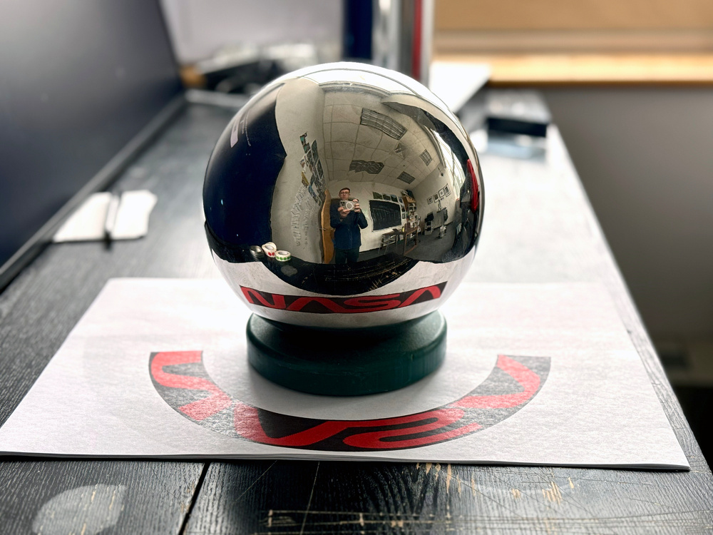 A reflective silver sphere rests on a table, capturing an upside-down reflection of its surroundings with a NASA logo visible beneath it.