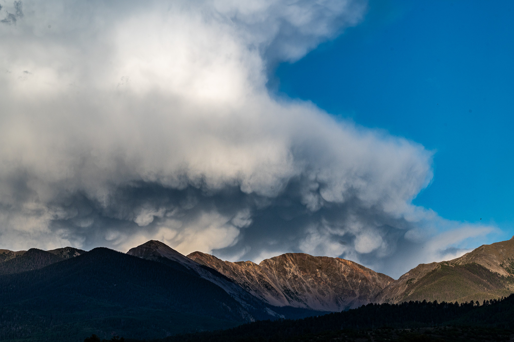 Dark thunderstorm over New Mexico's Truchas peaks, blue sky to right, dark mammatus clouds below, rising above top of frame. Mountains across the bottom third, some geologically rocky, others forested. A dramatic shot.