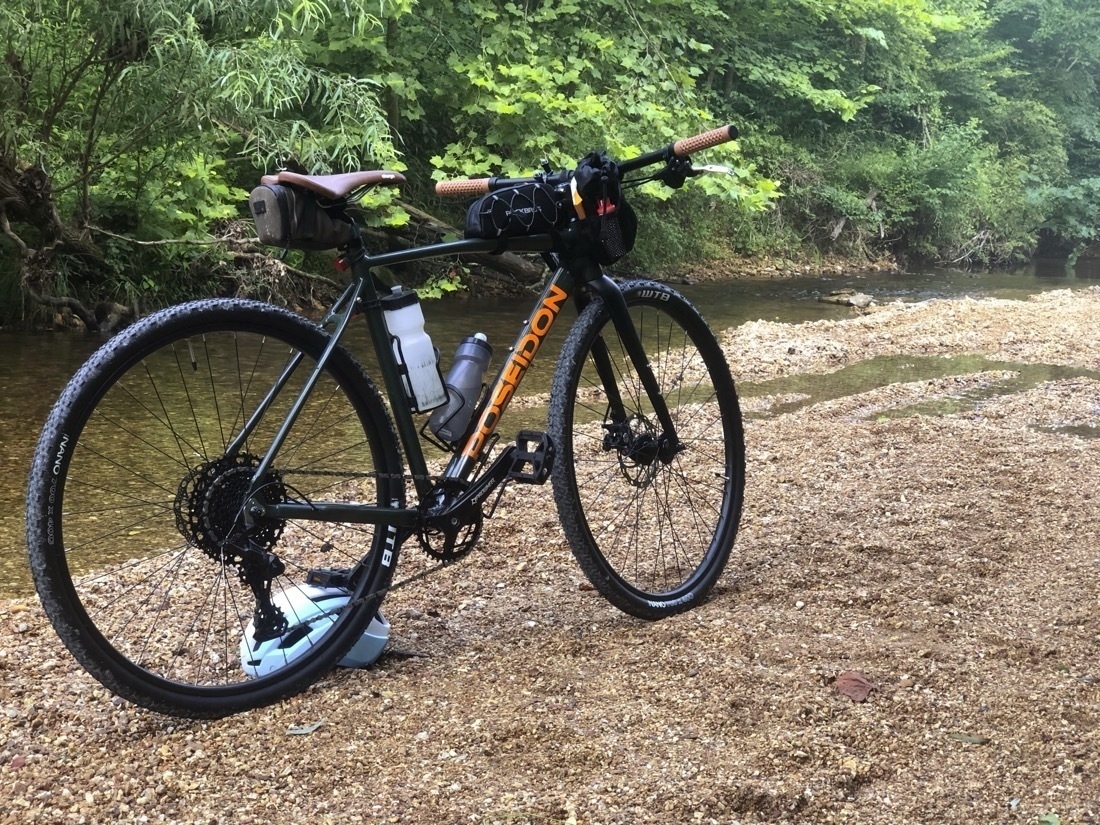 A green gravel style bike is propped up by a bike helmet on a gravel creek bed with a tree-lined creek in background.