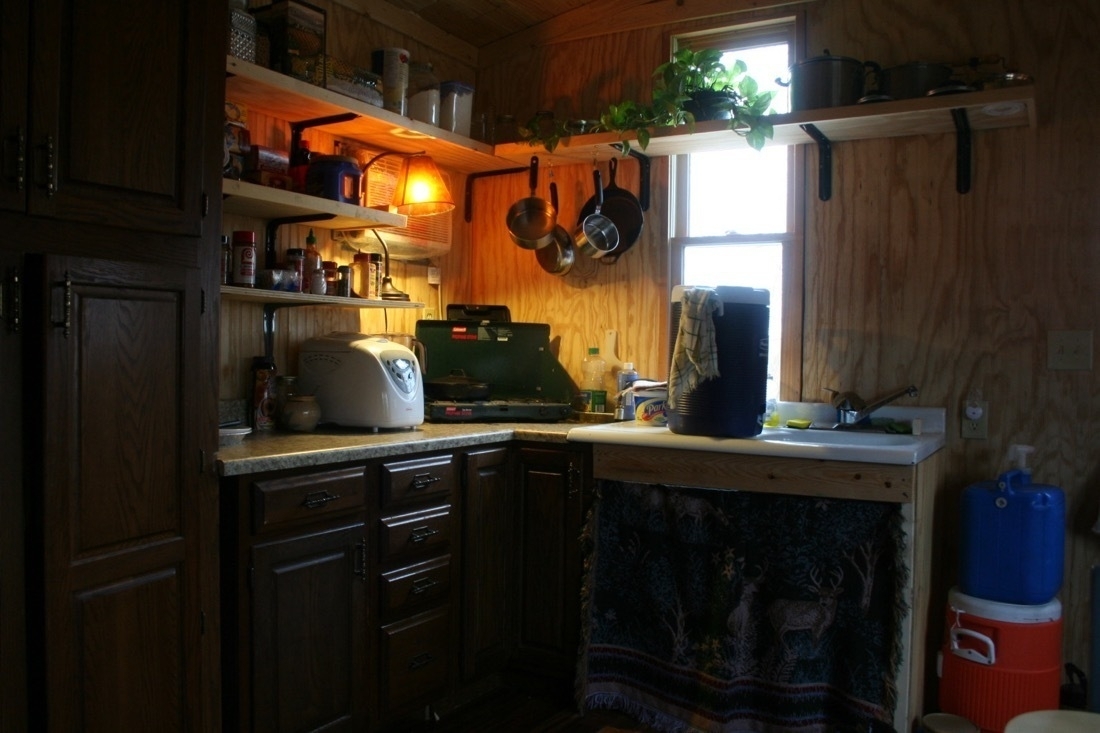 The interior of a tiny house kitchen area with a sink, water tanks, shelves, camping stove.