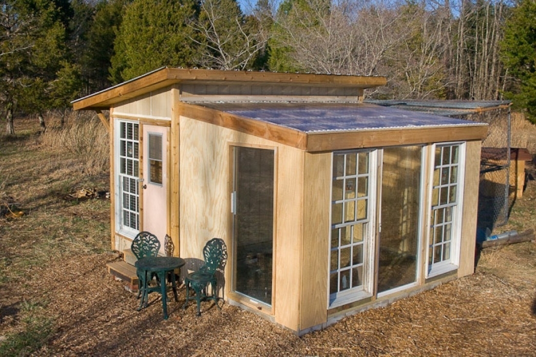 A small building with two halves. The further half is a chicken coop, the near side is a greenhouse. There is a small green table with chairs near the door of the greenhouse