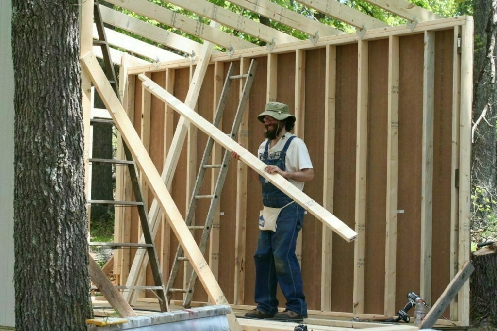 Bearded man carrying 2x4 lumber in construction of building with partial walls and partial roof