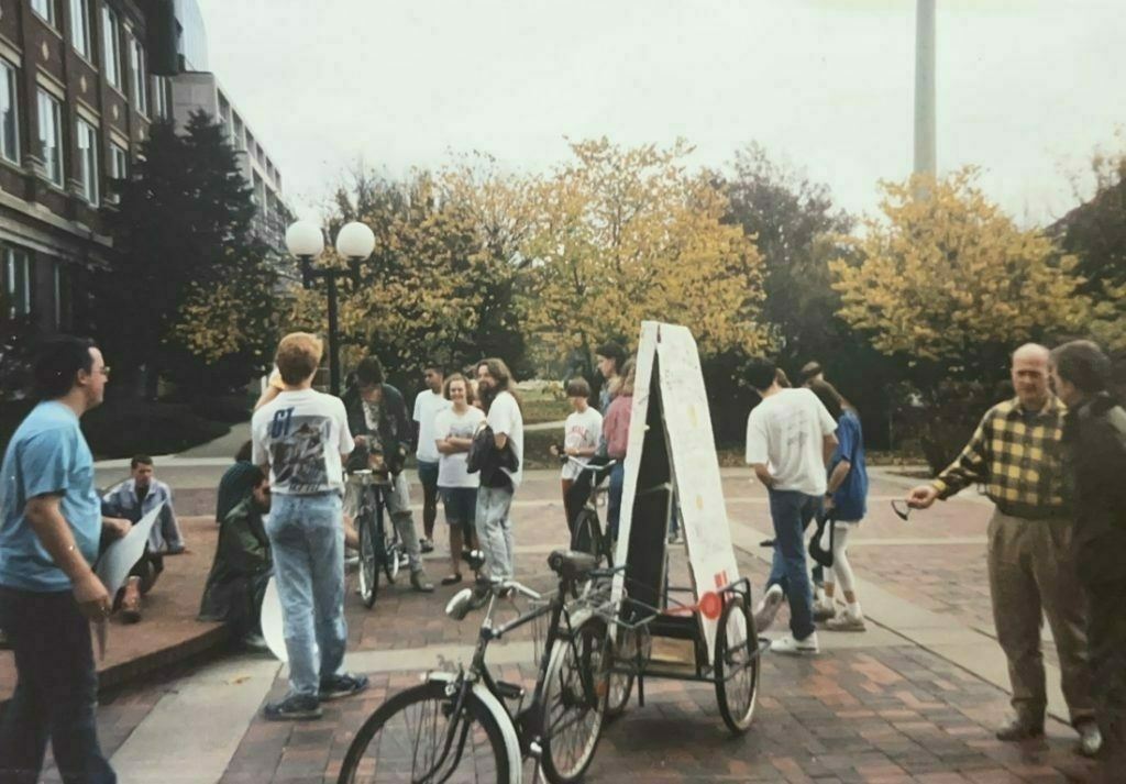 Small group of anti-war protesters gathered, holding signs. A bicycle is in center of image with sign in a small trailer attached.