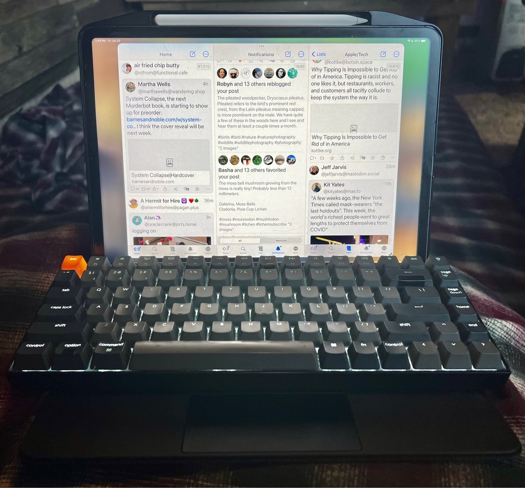 A mechanical Keychron keyboard rests in front of an iPad, set on top of the Magic Keyboard. On the screen is the Mona Mastodon app as I'm currently taking a break.
