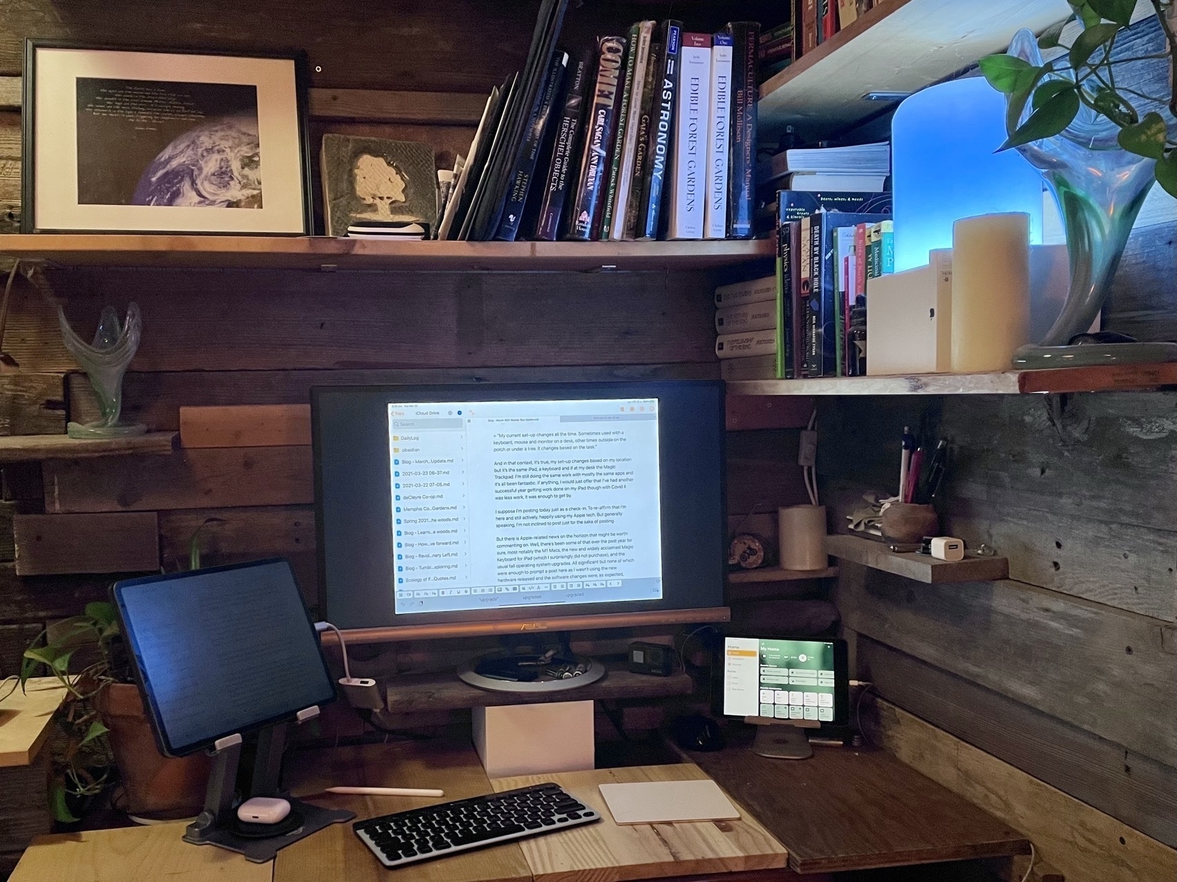 An iPad Pro is in a stand on a desk sitting to the lower left of an attached external monitor resting on a shelf. A smaller iPad sits to the lower right of the monitor.