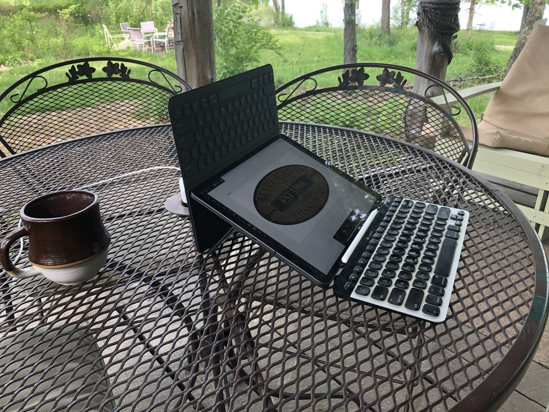 An iPad Pro in a Smart Keyboard Folio is turned upside down at an angle that is optimal for using an Apple Pencil. Affinity Designer is the app being used and an oval logo can be seen in the application window. The arrangement is on a metal table on a porch outside. A keyboard sits in front of the iPad.