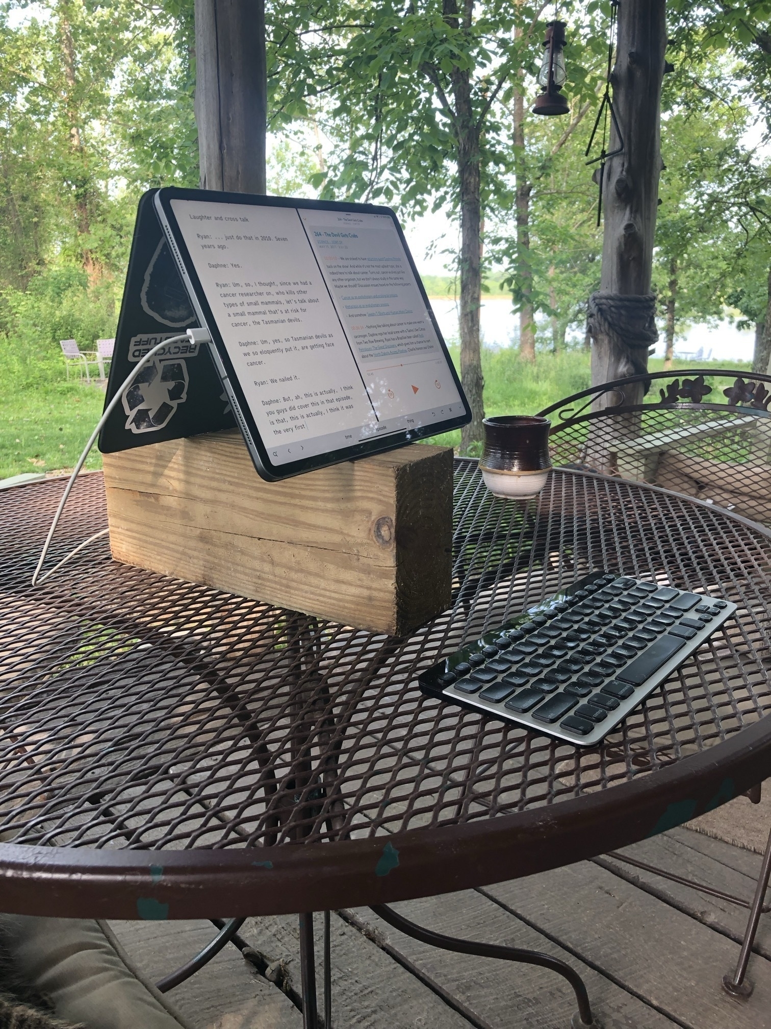 An iPad Pro in a Smart Keyboard Folio is arranged in a sort of A-Frame and is resting on a large wood block elevating it to eye height. The arrangement is an a metal table sitting on a porch. A keyboard sits in front of it.
