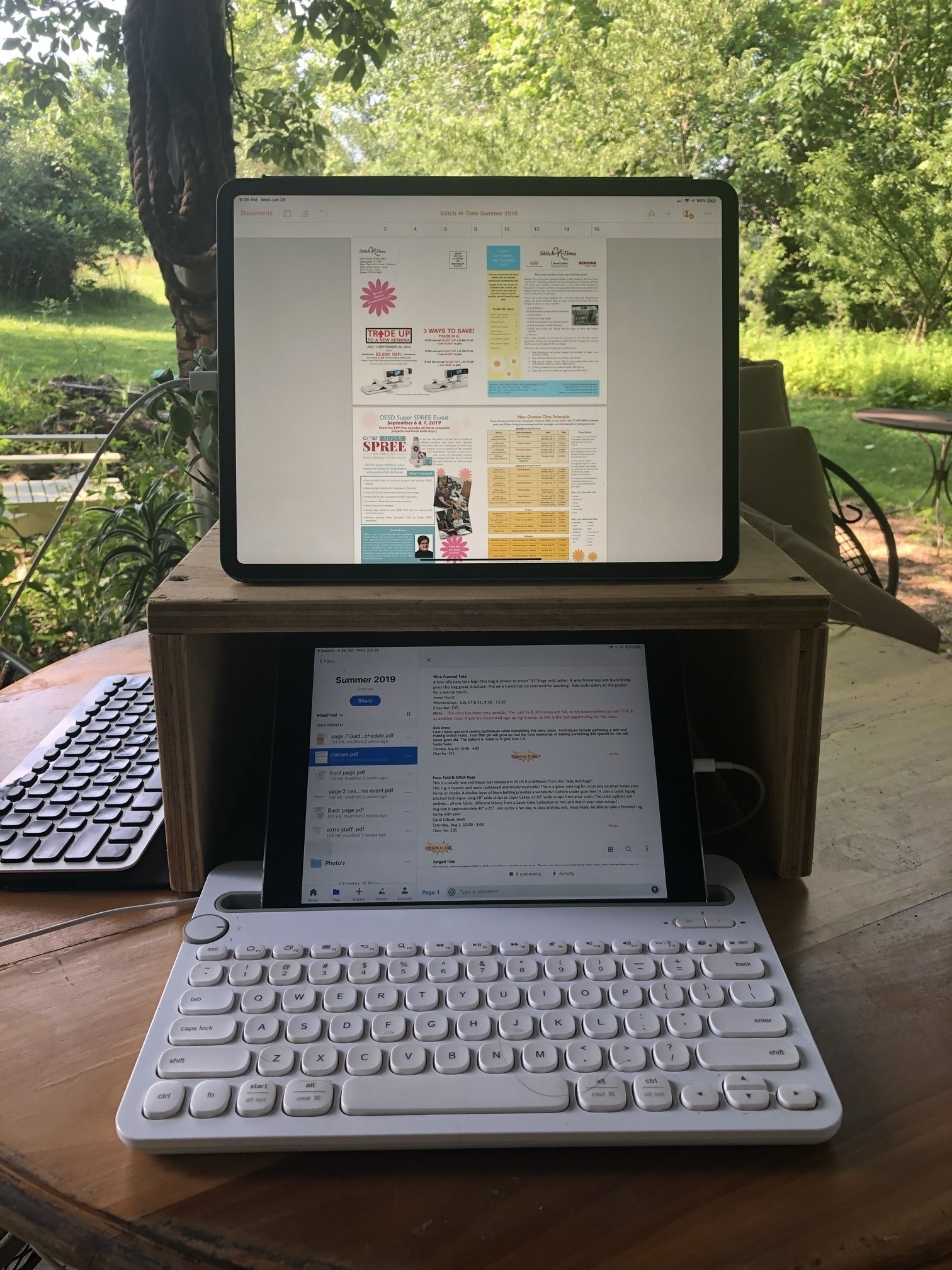 A small plywood box shelf is sitting on a table in the shade outside. Sitting on the table is a white keyboard that has a tablet slot holding an iPad. It's pushed up an inch inside the box shelf. Sitting up on top of the box is a larger iPad. The arrangement is being used as a sort of dual screen productivity set-up with the bottom iPad holding reference text which is being used to design a document on the iPad on the top of the shelf using the Pages application.
