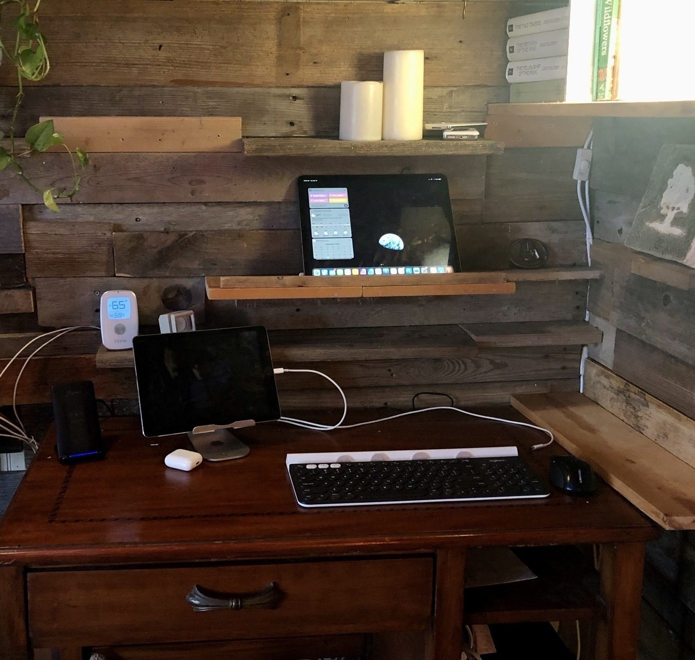 A desk near a wood plank wall with various shelves. An iPad is in a stand on the desk near a keyboard. A second iPad sits on a shelf at eye level behind the desk