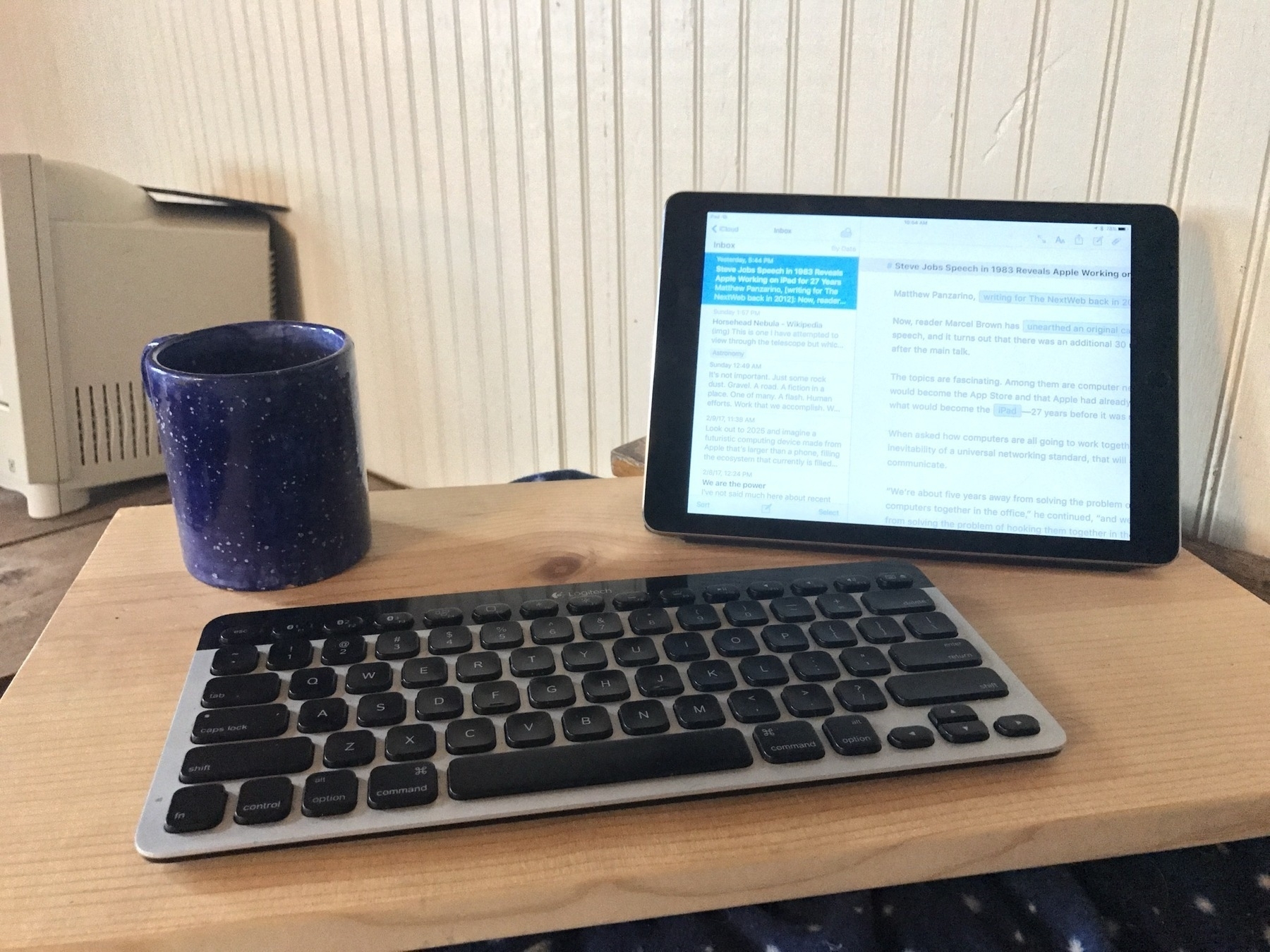 A plank of wood serves as an improvised lap desk. An iPad sits to the right, a blue coffee mug to the left and a keyboard in front.