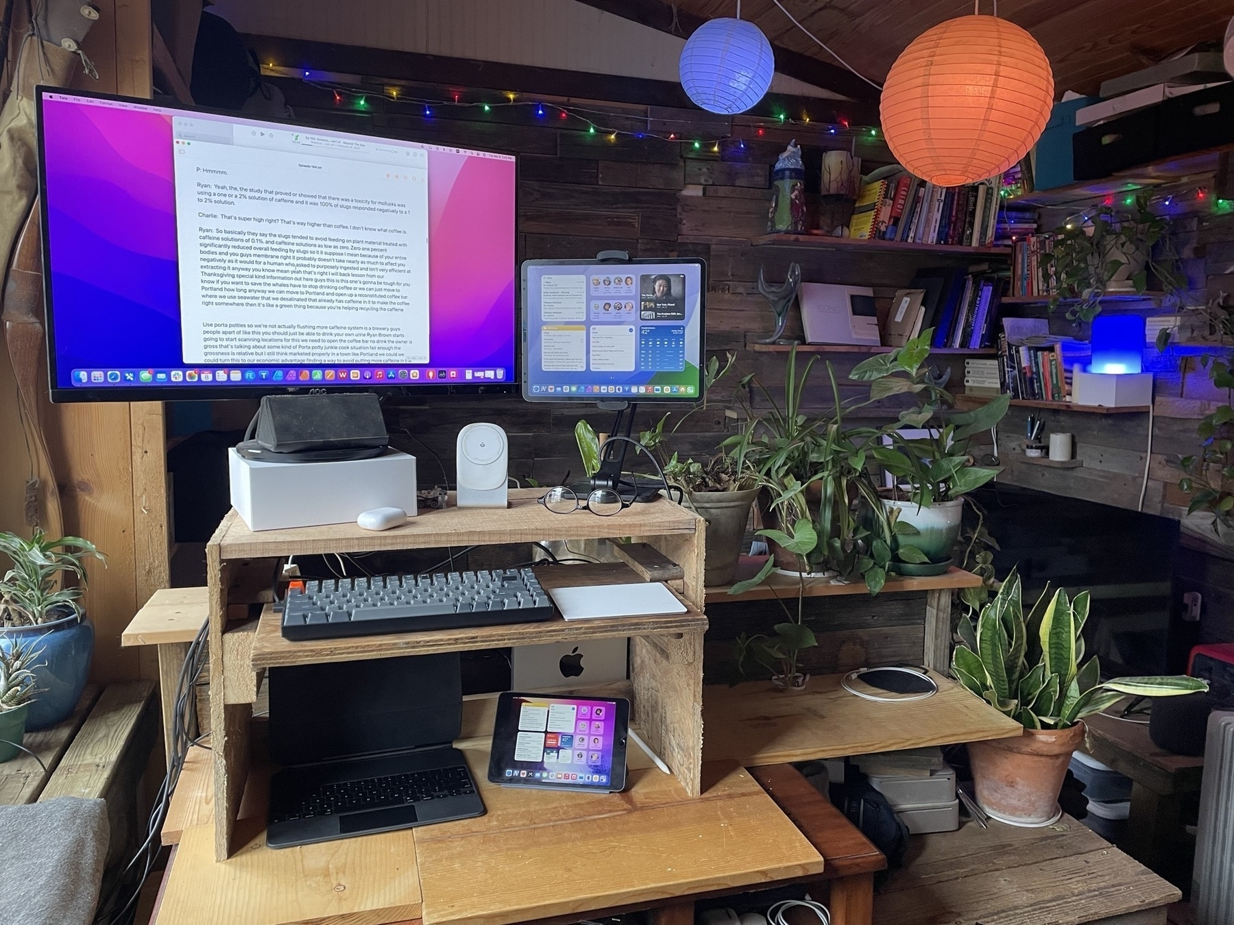 A standing desk has been improvised with a home built shelf on a desk. On the top shelf is a display attached to a Mac Mini. An iPad in a stand is to the right of the monitor. A keyboard and trackpad are on a second shelf below the top. Below that on the desk sits a smaller iPad.