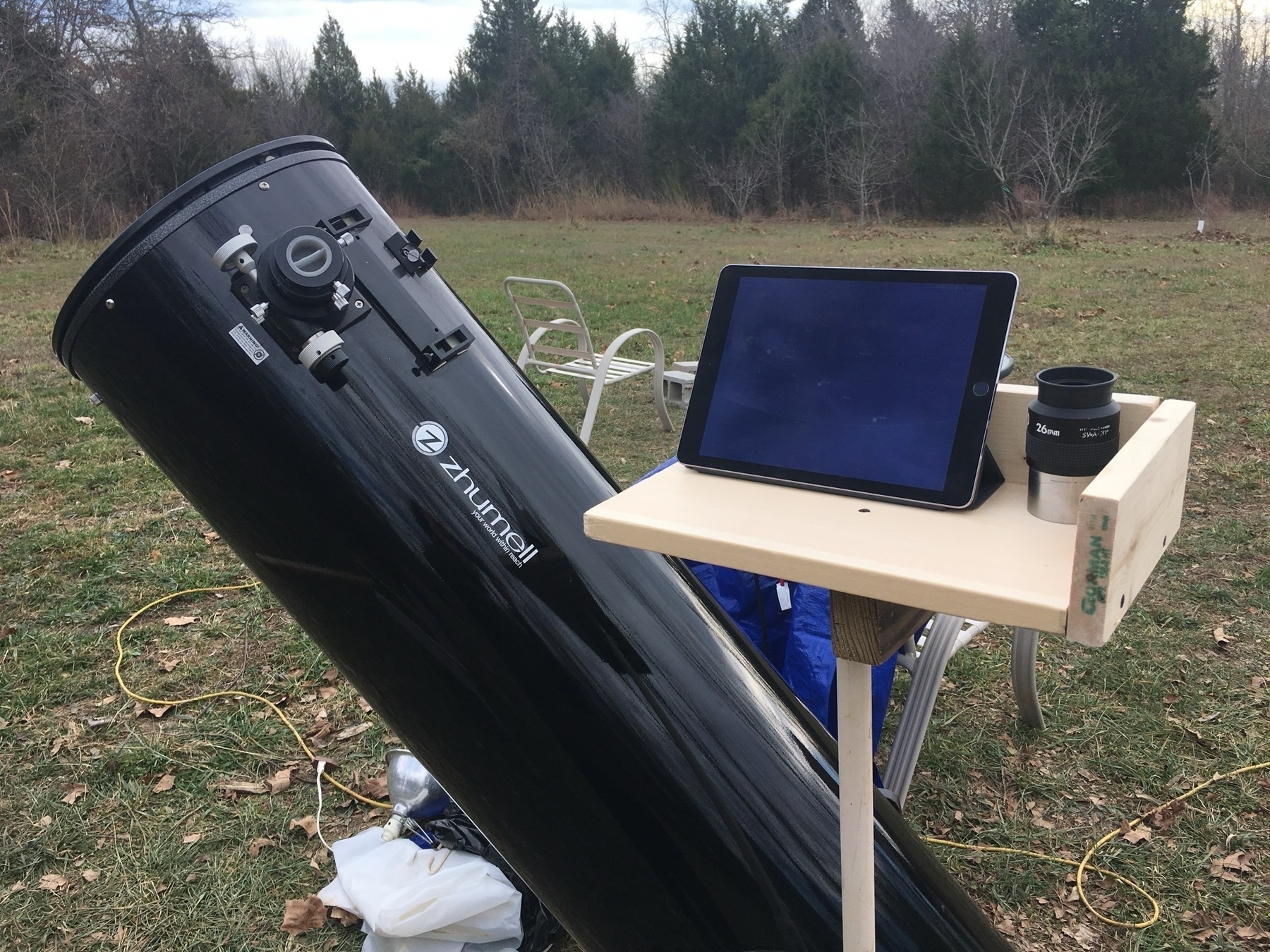 An iPad sits on a wood shelf that is attached to a large dobsonian telescope base which is below the bottom of photo, not visible. The wood plank is held up with a pipe. So that it is at standing level. The telescope is at an angle away from the iPad. 