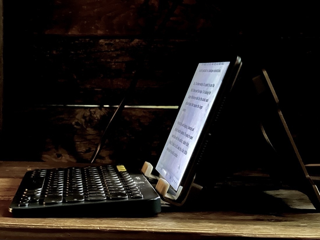 An iPad Mini sits in a stand on a wood shelf. The iPad is facing left, a black keyboard is in front of it. 