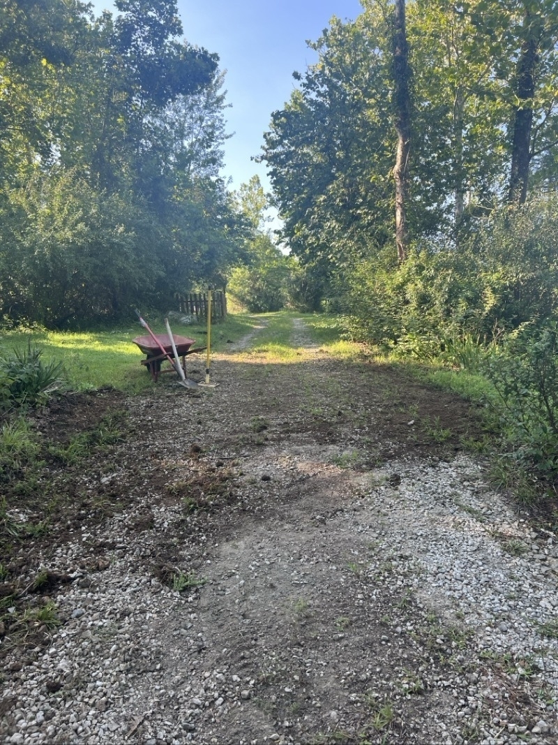 A gravel road looks a bit messy as dirt has been pulled across it to level it. A wheel barrow and tools are off to side of road.