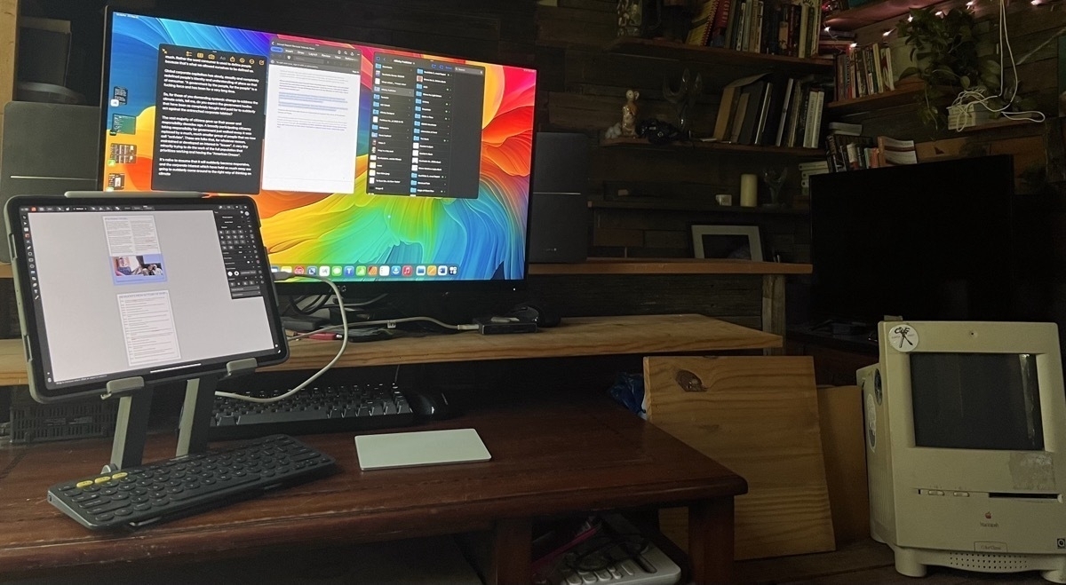 A iPad Pro in a stand sitting next to an external display. There is a keyboard and trackpad in front of the iPad and it is connected to the display.