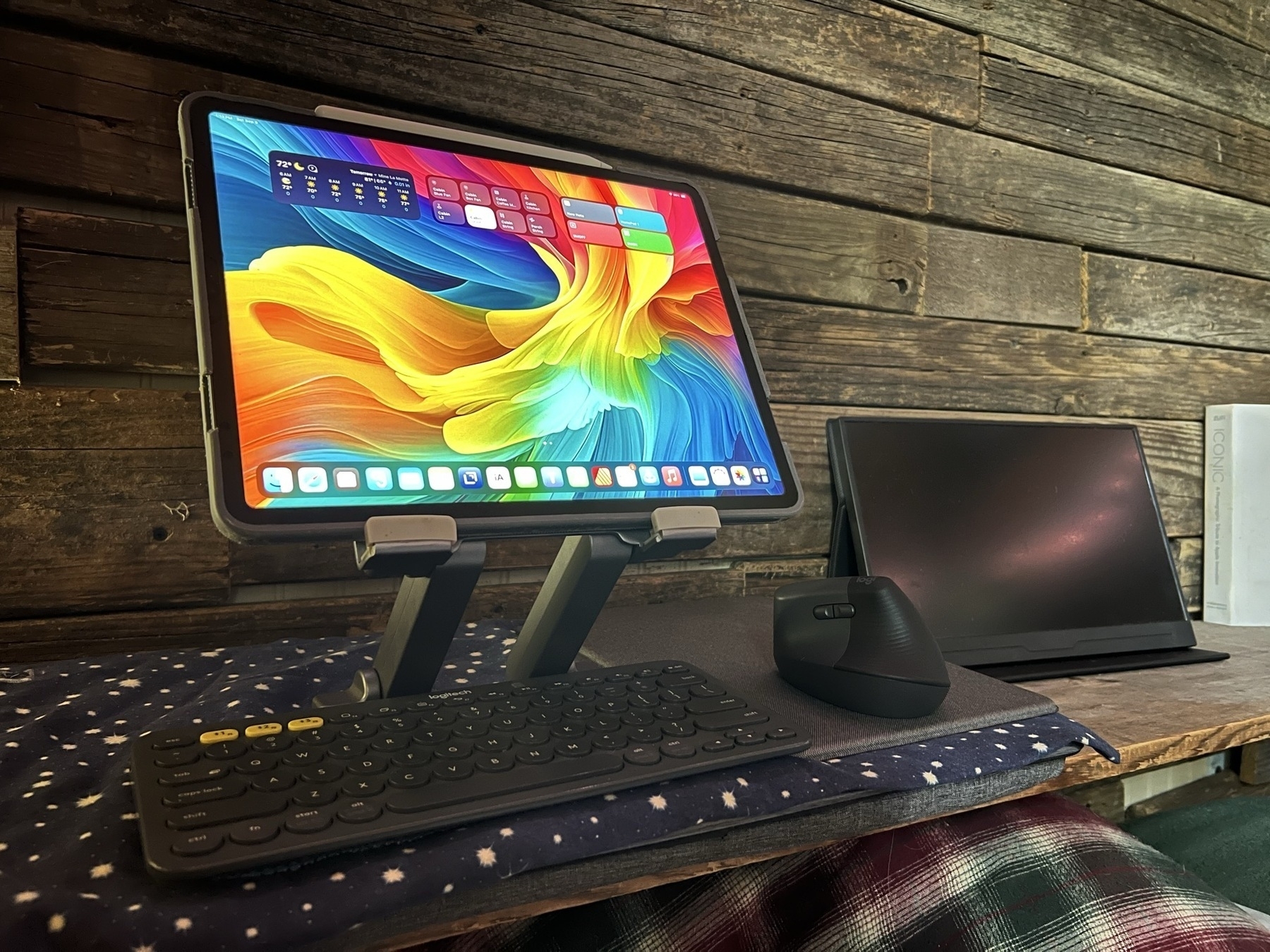 An iPad Pro is resting in a sturdy metal stand with a Logitech keyboard below it. To the right is a mouse. These are all resting an a lap desk which is sitting on a shelf next to a wood plank wall. 