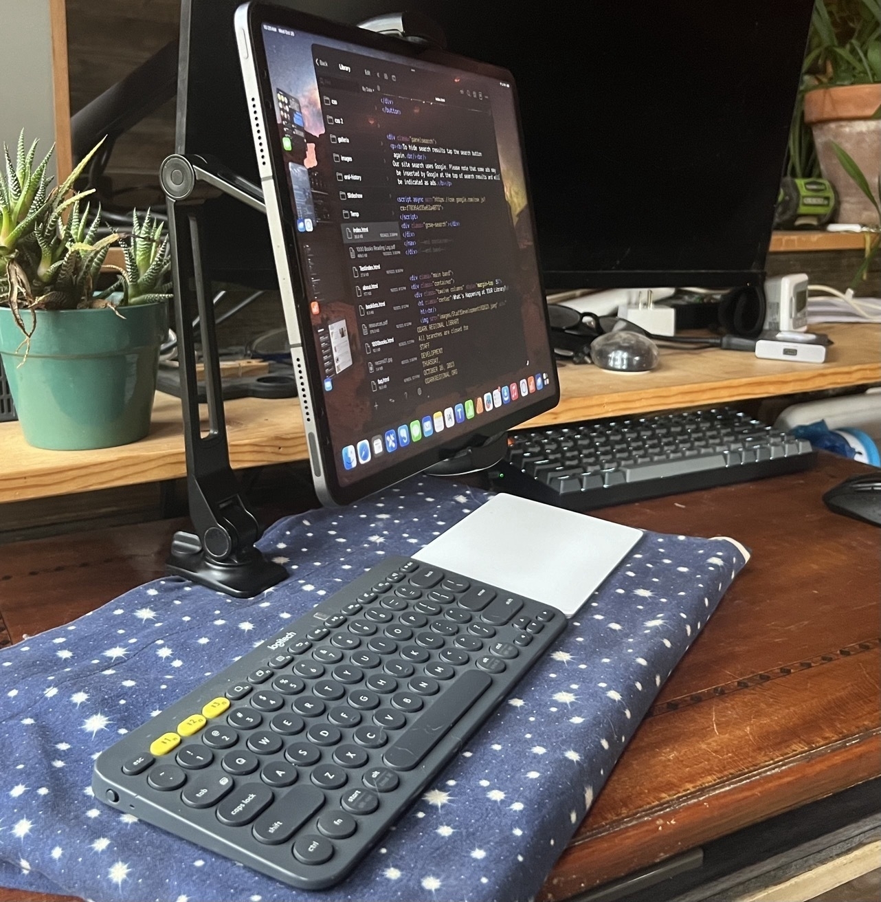 An iPad Pro floats above keyboard and trackpad on a lap desk. The iPad is held up by a configurable arm that is clamped to the back of the wood lap desk. Image taken from the side showing the arm.