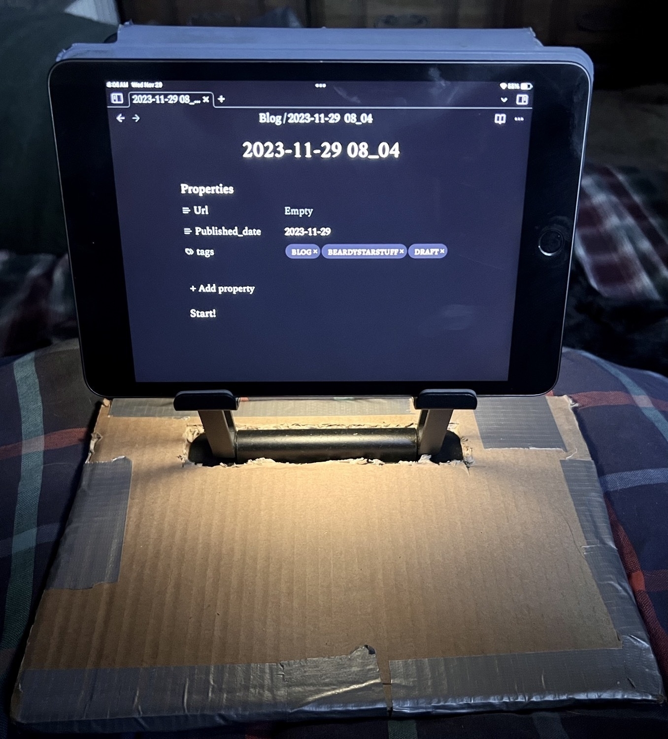 An iPad Mini  sits in a stand that has a cardboard base and is resting on a pillow. 