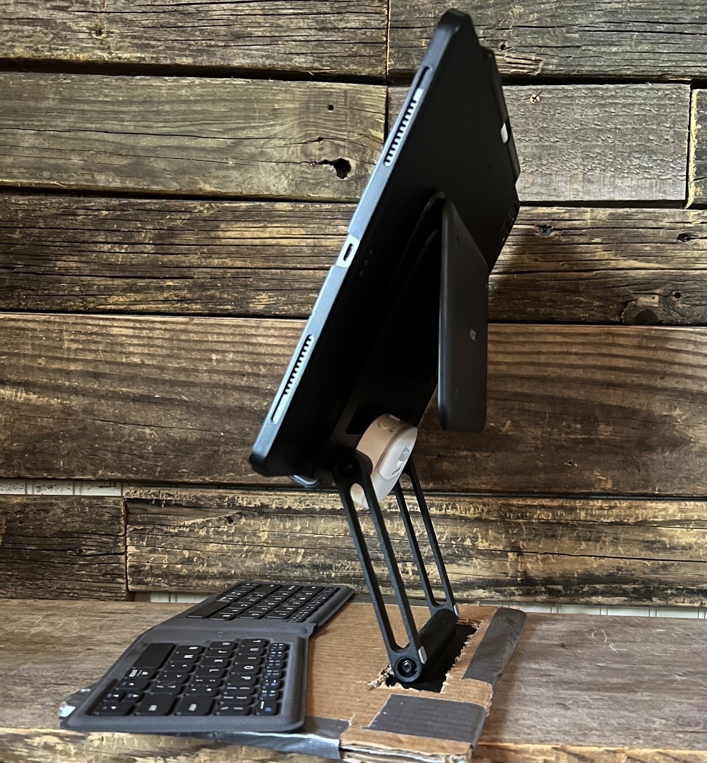 An iPad Pro  sits in a stand that has a cardboard base. The image is taken from the side, rear and shows that an attached, folded Moft stand is clamped over the back metal slap of the stand to hold the iPad in place 